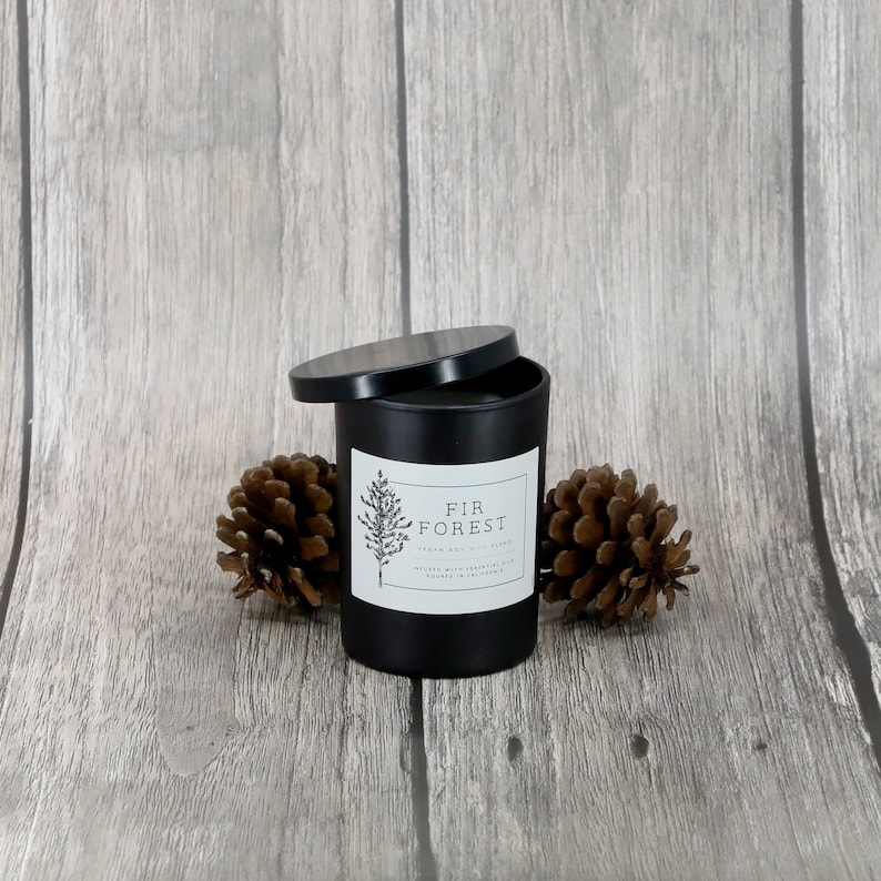 May include: Black candle jar with a white label that reads "FIR FOREST" and a small pine tree illustration. The candle is on a wood surface with two pine cones on either side.