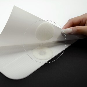 May include: Two white adhesive dots are attached to a white surface. The adhesive dots are being peeled off the surface.