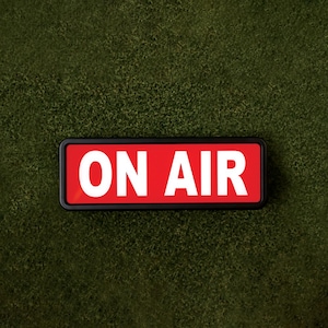 May include: A rectangular sign with a black frame and a bright red background featuring the text ON AIR in bold white capital letters. The sign is designed for broadcast notification and displays clear, high-contrast typography for visibility.
