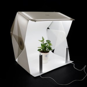 May include: A white foldable photo studio with two LED lights and a green plant in a white pot inside. The studio is set up on a black surface.