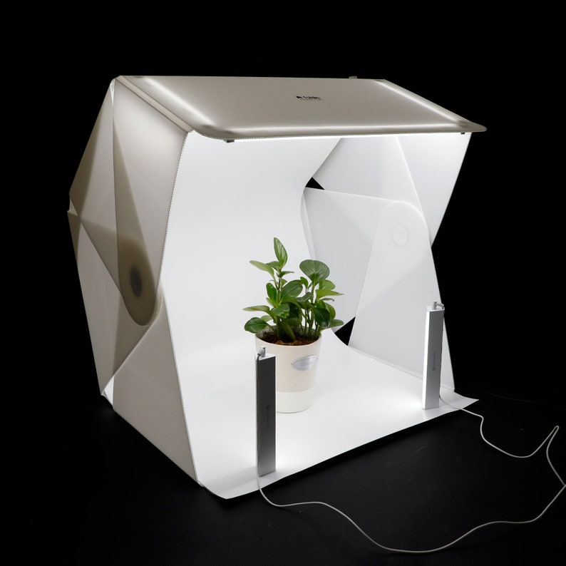 May include: A white foldable photo studio with two LED lights and a green plant in a white pot inside. The studio is set up on a black surface.