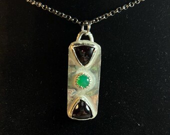 Puede incluir: Un collar con un colgante rectangular de plata con tres piedras preciosas. Las piedras preciosas de arriba y abajo son piedras triangulares negras, y la piedra preciosa del medio es una piedra redonda verde.