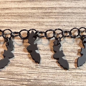 Può includere: Un braccialetto a catena nero con piccoli ciondoli a forma di pipistrello pendenti. Il braccialetto presenta una serie di maglie circolari collegate tra loro. Ogni ciondolo a forma di pipistrello è una silhouette nera. Il braccialetto è esposto su una superficie di legno.