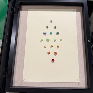 May include: A black shadow box frame containing a cream-colored rectangular mat. Small, polished, multi-colored stones are arranged in a vertical diamond pattern on the mat, featuring shades of purple, blue, green, yellow, orange, and red.