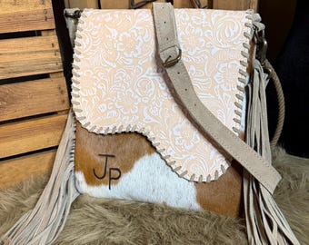 Puede incluir: Un bolso bandolera con una sección inferior de piel de vaca blanca y marrón con las letras Jp impresas en negro. La solapa superior es de color melocotón claro con un patrón floral blanco, acabado con costuras y largos flecos beige.