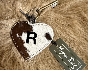 May include: A heart-shaped keychain with a cowhide pattern, featuring the letter "R" in black. The keychain is attached to a key and a tag that reads "Myra Bag". The background is a textured, brown surface.