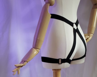 May include: A black adjustable harness with a metal ring. The harness is worn on a wooden mannequin.