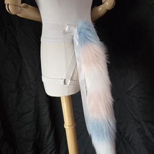 May include: A long, fluffy faux fur tail featuring a color-blocked pattern of light blue, soft pink, and white. The tail is attached to a white elastic belt with a white ribbon bow and includes a white elastic leg strap with a metal ring connector.
