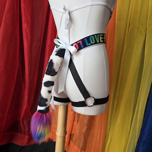 May include: A white harness featuring a black elastic waistband with the text "LOVE" in rainbow colors. A faux fur tail with a black and white cow print pattern and a multi-colored rainbow tip is attached to the back with a white ribbon bow.