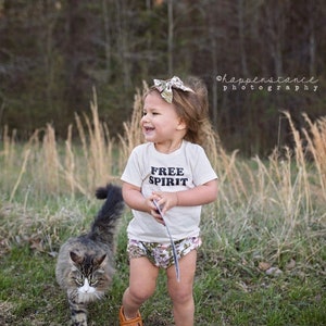 May include: A young child wearing a white t-shirt with the text "FREE SPIRIT" printed on it, brown shoes, and a floral print diaper cover. The child is standing in a grassy field with a cat walking in front of them.