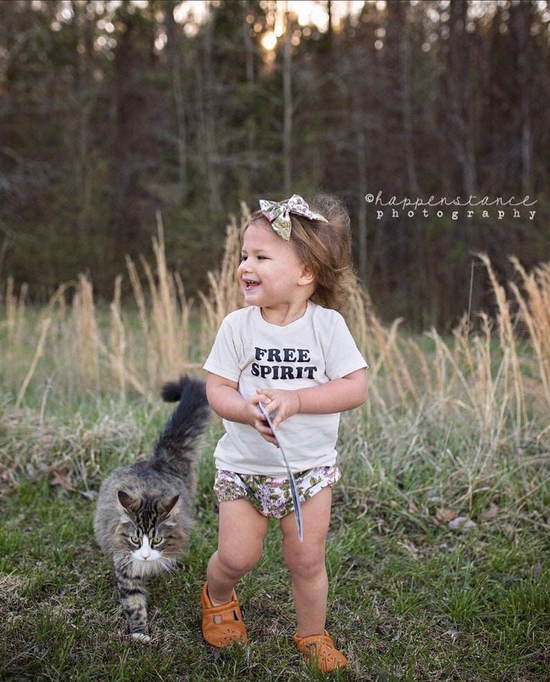 May include: A young child wearing a white t-shirt with the text "FREE SPIRIT" printed on it, brown shoes, and a floral print diaper cover. The child is standing in a grassy field with a cat walking in front of them.