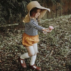 May include: A young girl wearing a striped long-sleeve shirt, mustard yellow bloomers, white knee-high socks, and brown shoes. She is holding a pink flower and wearing a wide-brimmed straw hat.