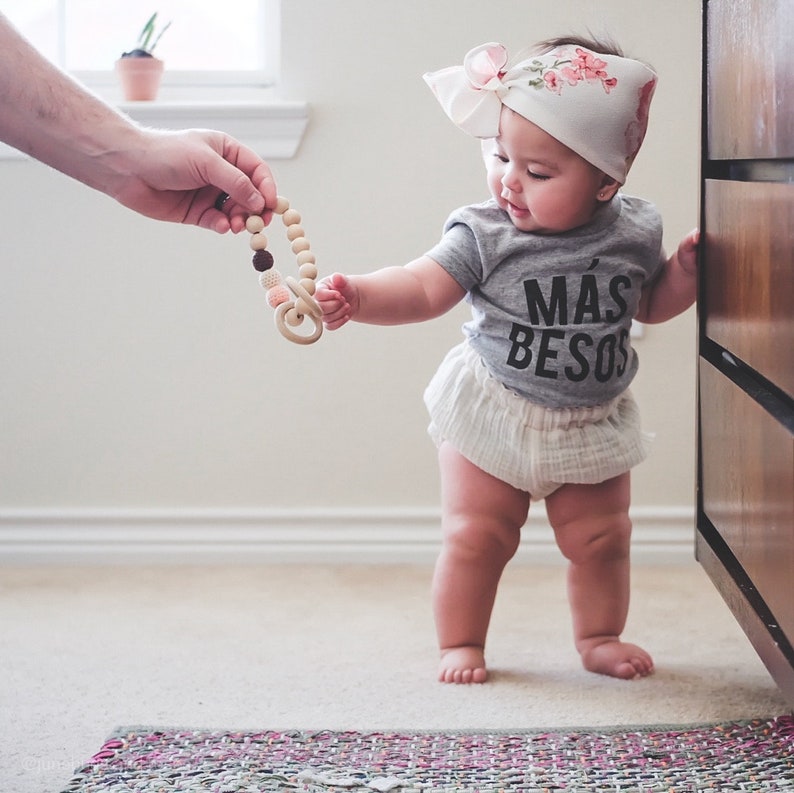 May include: A baby girl wearing a gray shirt with the text "M&aacute;s Besos" and a white floral headband is reaching for a wooden teething toy.