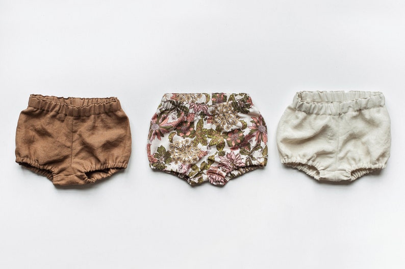 May include: Three pairs of baby bloomers in different colors and patterns. One pair is brown, one is white with a floral print, and one is white.