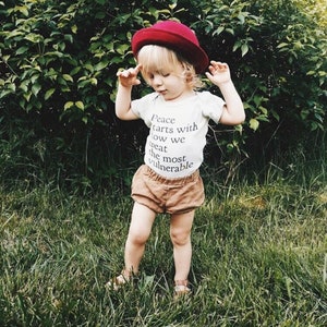 May include: A young child wearing a white t-shirt with the text "Peace starts with how we treat the most vulnerable" printed on it. The child is also wearing a red hat and brown shorts. The child is standing in a grassy area.