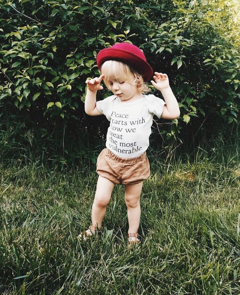 May include: A young child wearing a white t-shirt with the text "Peace starts with how we treat the most vulnerable" printed on it. The child is also wearing a red hat and brown shorts. The child is standing in a grassy area.