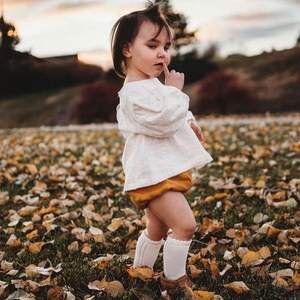 May include: A young child wearing a white blouse, a mustard yellow skirt, and white knee-high socks with lace detailing. The child is standing in a field of fallen leaves.