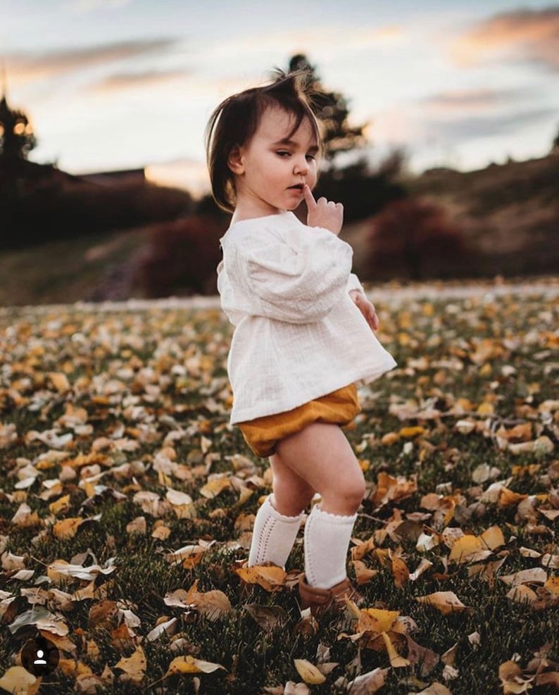 May include: A young child wearing a white blouse, a mustard yellow skirt, and white knee-high socks with lace detailing. The child is standing in a field of fallen leaves.