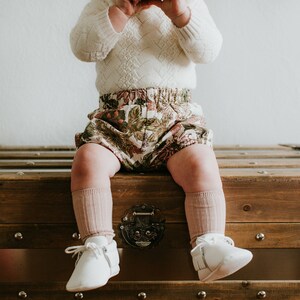 May include: A baby wearing a white sweater, floral print bloomers, and pink knee-high socks sits on a wooden chest. The baby is wearing white shoes.