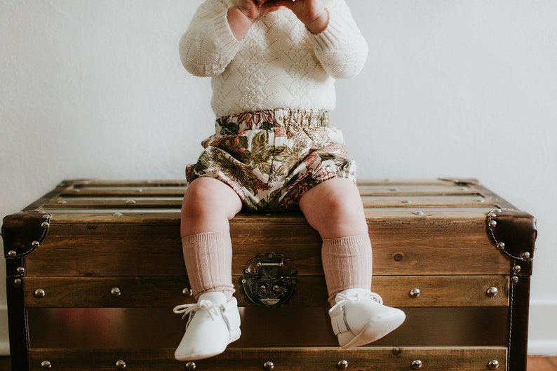 May include: A baby wearing a white sweater, floral print bloomers, and pink knee-high socks sits on a wooden chest. The baby is wearing white shoes.