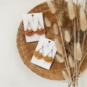 May include: Two pairs of handmade macrame earrings in terracotta and mustard yellow, displayed on white cards with the text "Feather + Fringe". The earrings are set against a woven basket and dried floral arrangement.