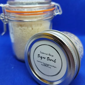 May include: Two glass jars of sugar scrub on a blue background. One jar is upright, displaying the contents, while the other is on its side, revealing a label that reads "Essential Reign Sugar Scrub." The ingredients listed are granulated cane sugar, argon oil, and essential oil of lime.