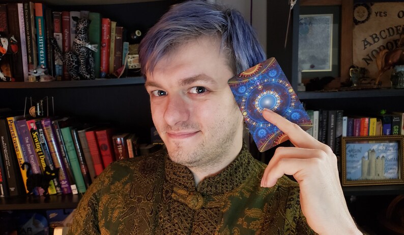Puede incluir: Una persona con cabello azul sostiene una carta de tarot con un dise&ntilde;o de mandala azul y dorado. La carta tiene un c&iacute;rculo blanco en el centro con un punto azul en el medio. La persona lleva una camisa con estampado verde y dorado.