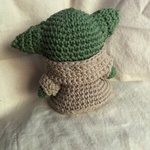 May include: A handmade crochet figure of a character with large ears, wearing a beige robe. The figure's head and ears are green, and the robe is a light beige colour. The figure is sitting on a white surface.