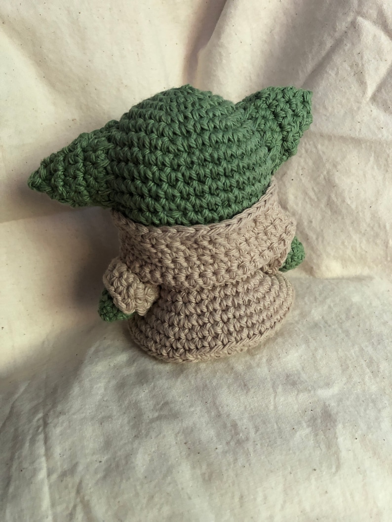 May include: A handmade crochet figure of a character with large ears, wearing a beige robe. The figure's head and ears are green, and the robe is a light beige colour. The figure is sitting on a white surface.