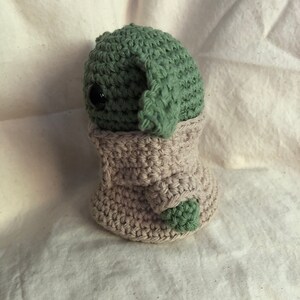 May include: A handmade crochet doll of a character with large ears and a beige robe. The doll's head and ears are green, and it has a black eye. The robe is beige, and the doll is on a white surface.
