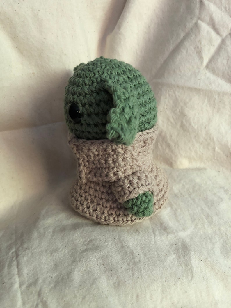 May include: A handmade crochet doll of a character with large ears and a beige robe. The doll's head and ears are green, and it has a black eye. The robe is beige, and the doll is on a white surface.