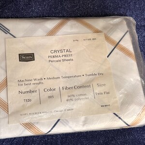 Puede incluir: Un juego empaquetado de sábanas de percal Sears Crystal Perma-Prest. Las sábanas blancas tienen un patrón geométrico en oro y azul marino. El embalaje incluye una etiqueta con los detalles del producto, incluido el contenido de fibra y el tamaño.