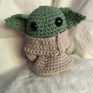May include: Handmade crochet figure of a character with large ears, green skin, and black eyes. The figure is wearing a beige robe. A collectible or toy.