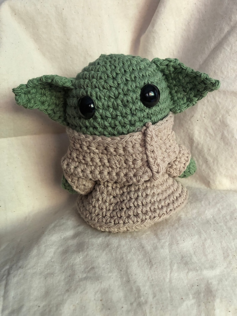 May include: Handmade crochet figure of a character with large ears, green skin, and black eyes. The figure is wearing a beige robe. A collectible or toy.
