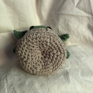 May include: A handmade crochet turtle toy. The turtle has a light brown body, green head and flippers, and black eyes. The toy is made with a tight crochet stitch, giving it a textured appearance. The turtle is lying on a white surface.