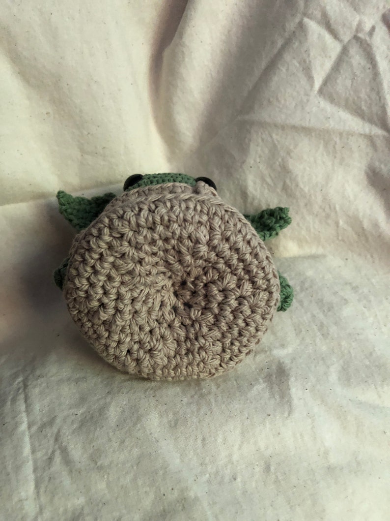 May include: A handmade crochet turtle toy. The turtle has a light brown body, green head and flippers, and black eyes. The toy is made with a tight crochet stitch, giving it a textured appearance. The turtle is lying on a white surface.