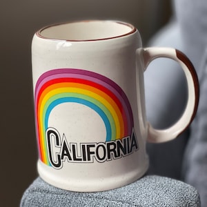 Pode incluir: Caneca de cerâmica branca com um design de arco-íris e a palavra "CALIFORNIA" em letras pretas. A caneca tem uma borda e uma alça marrons.