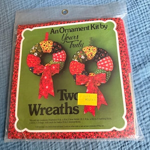 May include: An ornament kit by Yours Truly, featuring two wreaths. The wreaths are made of colorful fabric patches and adorned with red bows. The kit includes fabric, rings, rick rack, and instructions. The package is sealed in a clear plastic bag.