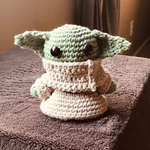 May include: A handmade crochet doll of a character with large ears and black eyes. The doll is primarily light green and beige, with a textured appearance. It is wearing a beige robe. The doll is sitting on a brown surface.