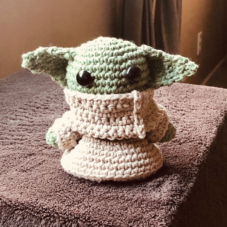 May include: A handmade crochet doll of a character with large ears and black eyes. The doll is primarily light green and beige, with a textured appearance. It is wearing a beige robe. The doll is sitting on a brown surface.