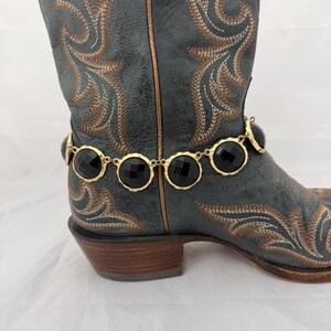 Op de afbeelding: Een close-up van een grijze cowboylaars met bruin en gouden borduursel. Een gouden ketting met zwarte cirkelvormige edelstenen is om de enkel van de laars gewikkeld. De laars heeft een spitse neus en een bruine hak.