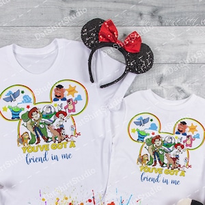 Może przedstawiać: Dwie białe koszulki z kolorowym wzorem głowy Myszki Miki przedstawiającym postacie z filmowej serii Toy Story. Wz&oacute;r zawiera Woody'ego, Buzz Lightyeara, Jessie i inne postacie. Tekst na koszulkach brzmi "You've got a friend in me".