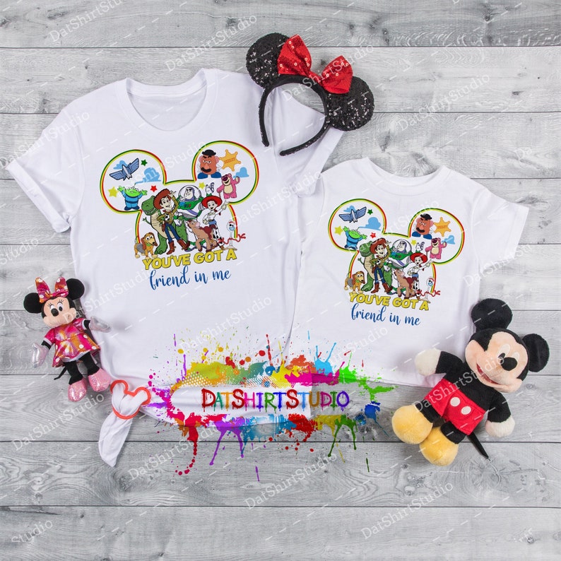 Może przedstawiać: Dwie białe koszulki z kolorowym wzorem głowy Myszki Miki przedstawiającym postacie z filmowej serii Toy Story. Wz&oacute;r zawiera Woody'ego, Buzz Lightyeara, Jessie i inne postacie. Tekst na koszulkach brzmi "You've got a friend in me".