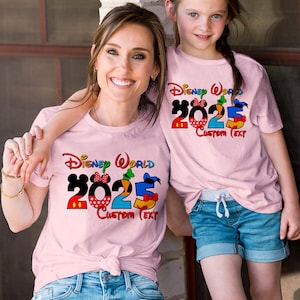 Może przedstawiać: Dwie r&oacute;żowe koszulki z napisem "Disney World 2023 Custom Text" w kolorowych literach. Koszulki przedstawiają postacie Myszki Miki, Minnie Mouse, Kaczora Donalda, Goofy'ego i Pluto.