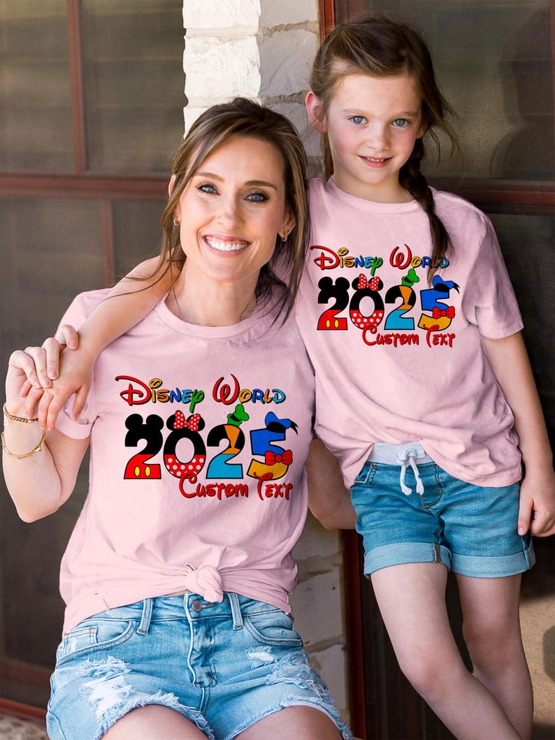 Może przedstawiać: Dwie r&oacute;żowe koszulki z napisem "Disney World 2023 Custom Text" w kolorowych literach. Koszulki przedstawiają postacie Myszki Miki, Minnie Mouse, Kaczora Donalda, Goofy'ego i Pluto.