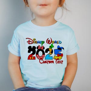 Może przedstawiać: Jasnoniebieska koszulka z kolorową grafiką Disney World 2025 przedstawiającą Myszki Miki, Minnie, Donalda i Goofy. Pod grafiką znajduje się tekst "Custom Text".