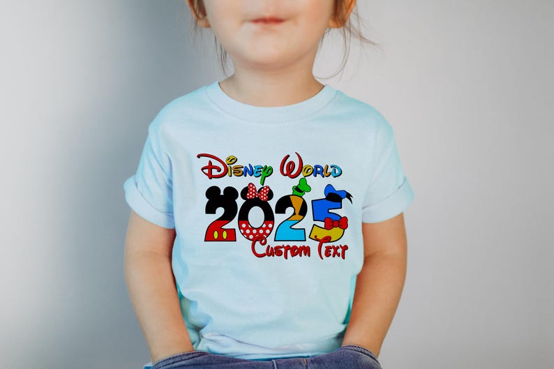 Może przedstawiać: Jasnoniebieska koszulka z kolorową grafiką Disney World 2025 przedstawiającą Myszki Miki, Minnie, Donalda i Goofy. Pod grafiką znajduje się tekst "Custom Text".