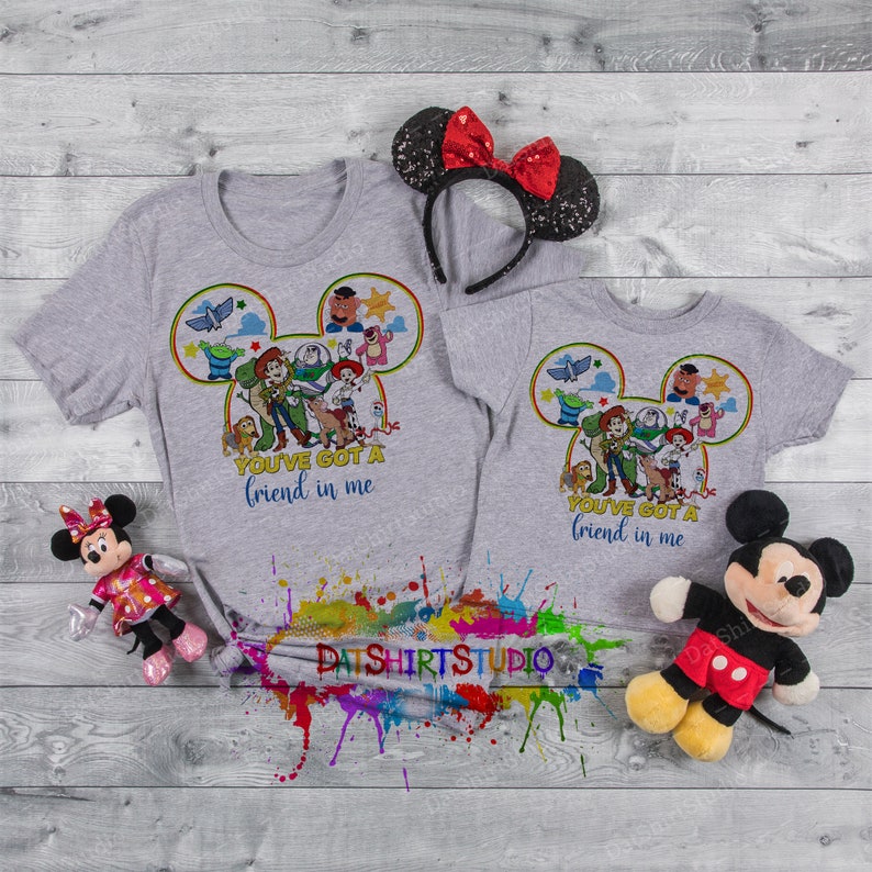 Może przedstawiać: Dwie szare koszulki z nadrukiem głowy Myszki Miki przedstawiającej postacie z serii film&oacute;w Toy Story. Tekst na koszulkach brzmi "You've got a friend in me".