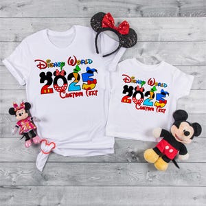 Może przedstawiać: Dwie białe koszulki z nadrukiem Disney World 2025. Nadruk przedstawia Myszke Miki, Minnie Mouse, Donalda Kaczora, Goofy'ego i Pluto. Na koszulkach widnieje tekst "Disney World 2025 Custom Text".