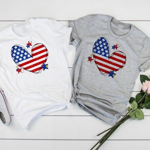 May include: Two t-shirts with a red, white, and blue heart-shaped American flag design. The shirts are white and grey and have a distressed look.